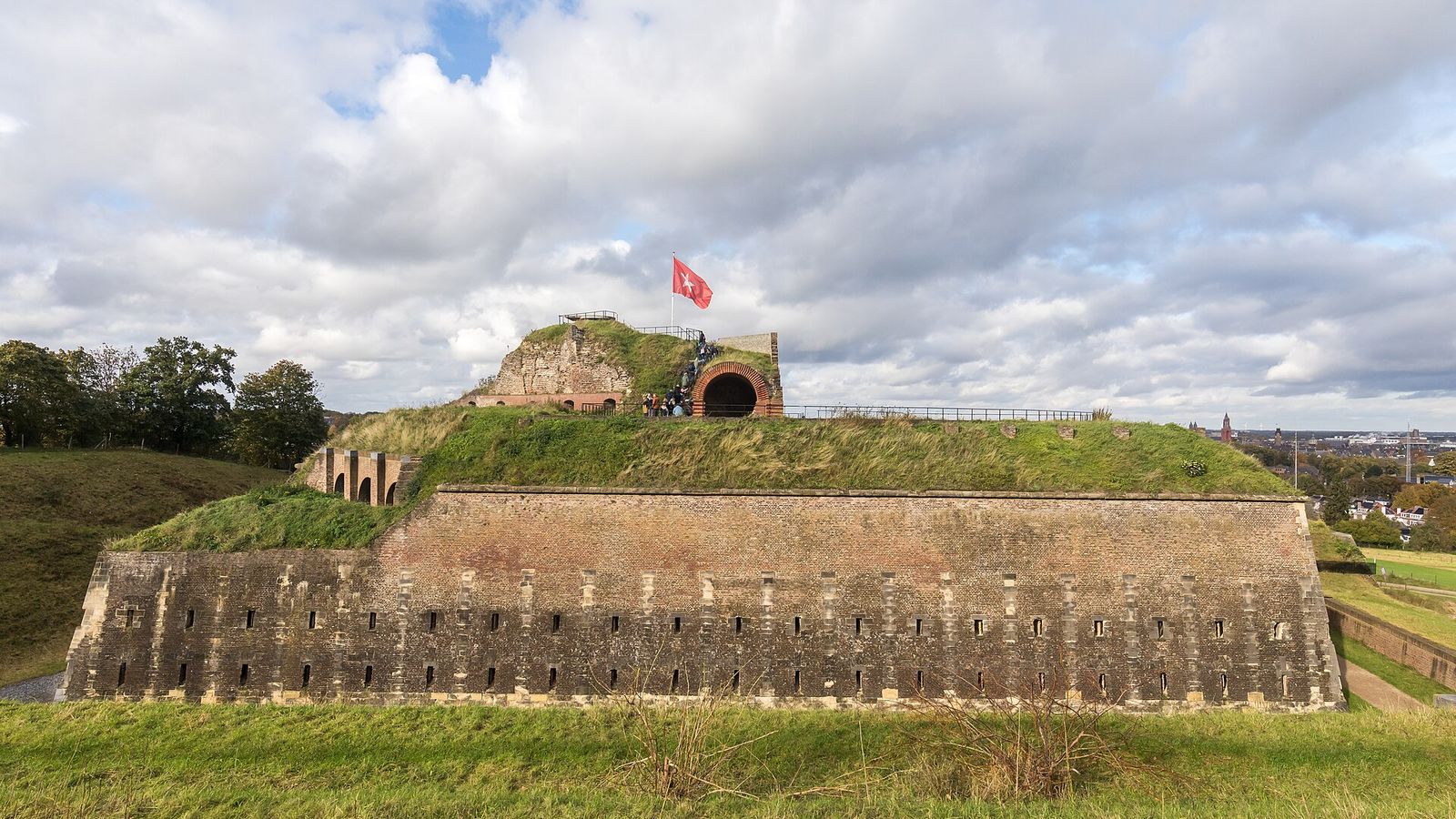 Fort Sint Pieter van opzij: een bakstenen muur met schietgaten onder een grasdak, bovenop een rode vlag met een witte ster