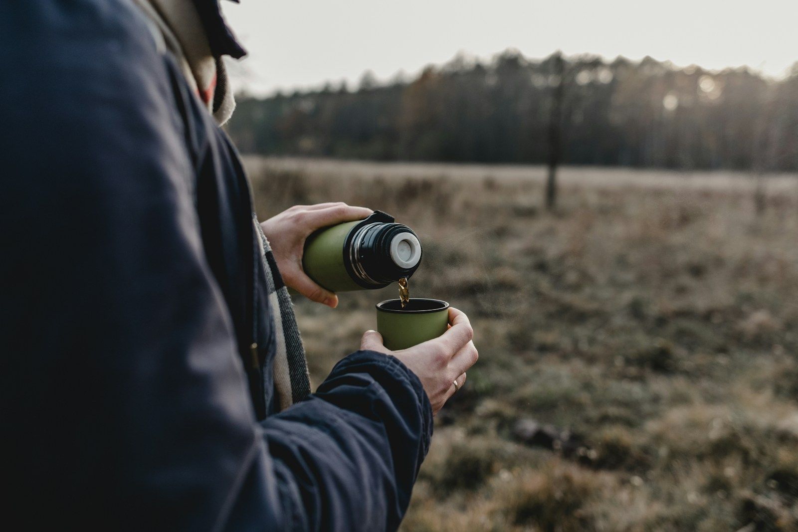 Twee handen schenken uit een groene thermosfles in een groene beker; de persoon draagt een donkerblauwe jas en een geruite sjaal, op de achtergrond een open veld met een bosrand