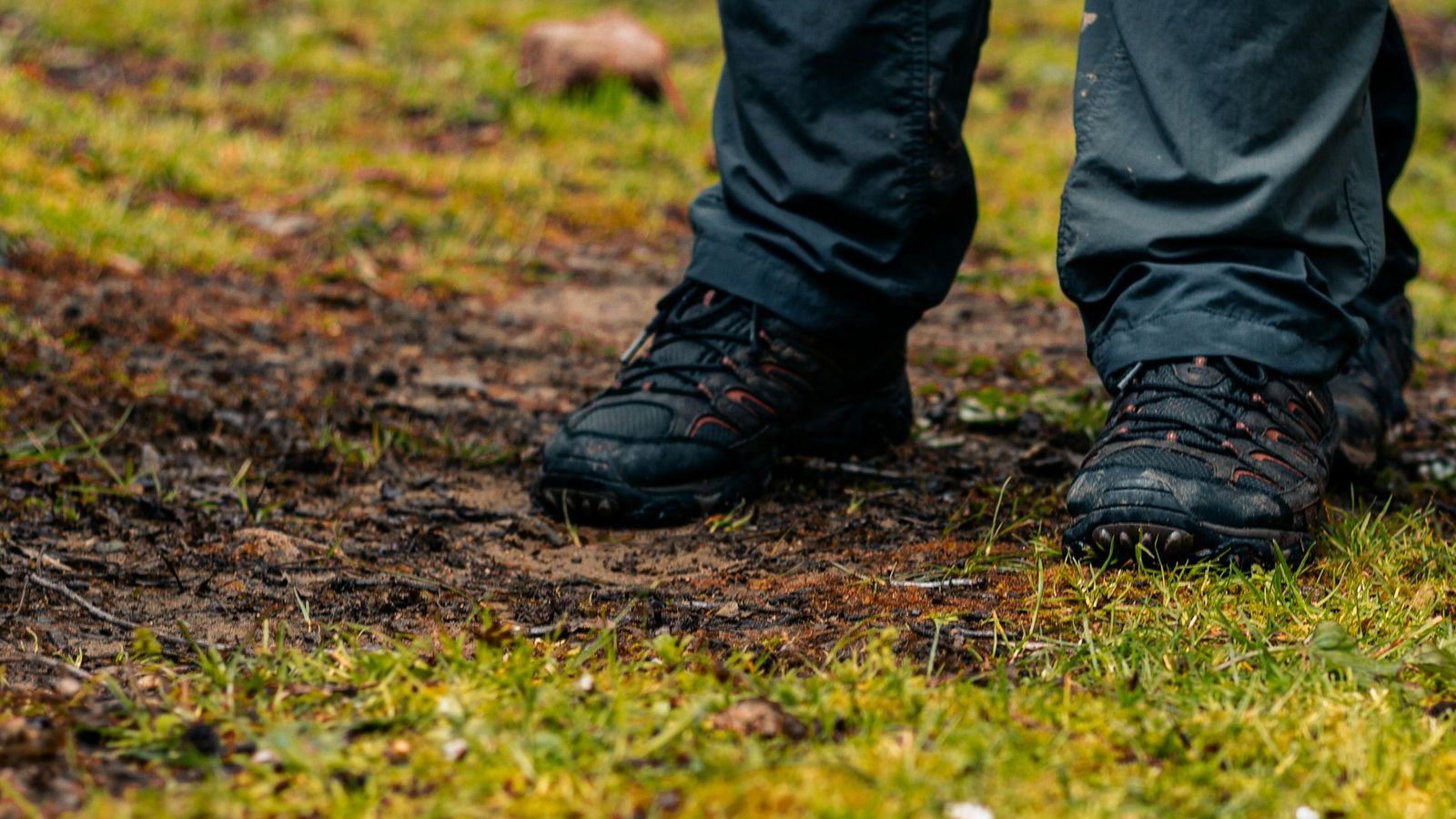 Onderbenen van een wandelaar op een nat grasveld met een modderige plek; donkere wandelschoenen met modder op de neus, de broekspijpen vallen over de enkel